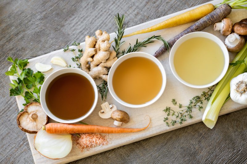 vegetables, herbs, and three bowls of broth sitting on a cutting board.