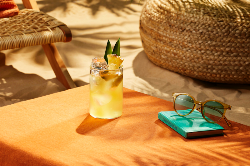 A glass of Life's a Beach cocktail beside a book and sunglasses on a beach table.