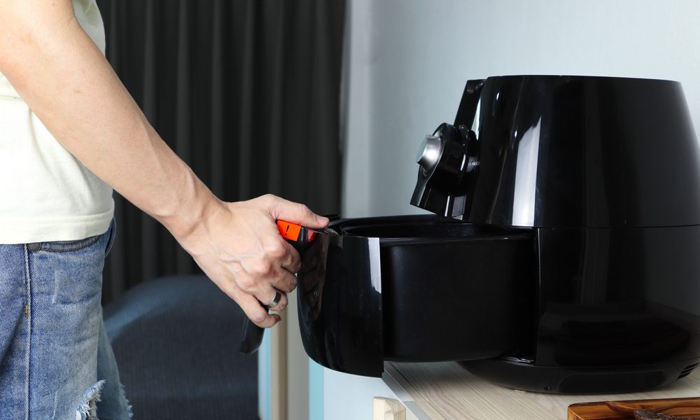 Right hand of man holding the tray of the black deep air fryer which is on the wooden table in the white kitchen.