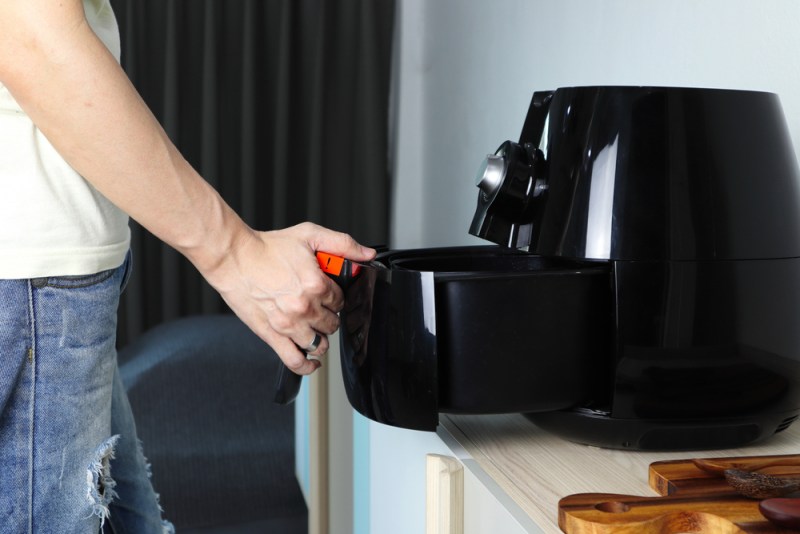 Right hand of man holding the tray of the black deep air fryer which is on the wooden table in the white kitchen.