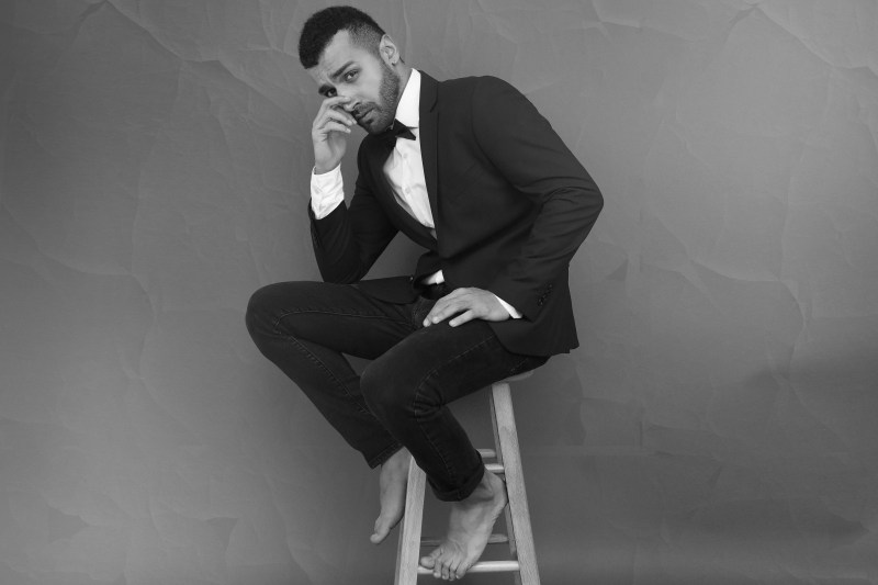 Barefoot man in a tuxedo sitting on a wooden stool.