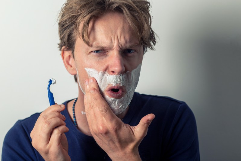 man with shaving cream on his face using a blue razor to shave.