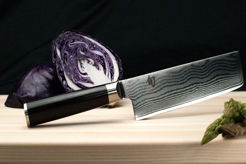A Shun knife with a sliced red cabbage behind it on a wooden cutting board.