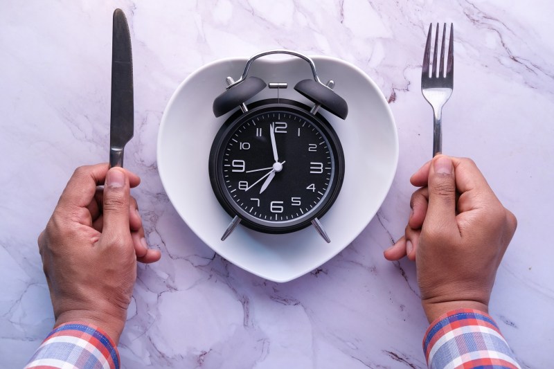 Man holding spoon and fork in between a clock placed on a plate.