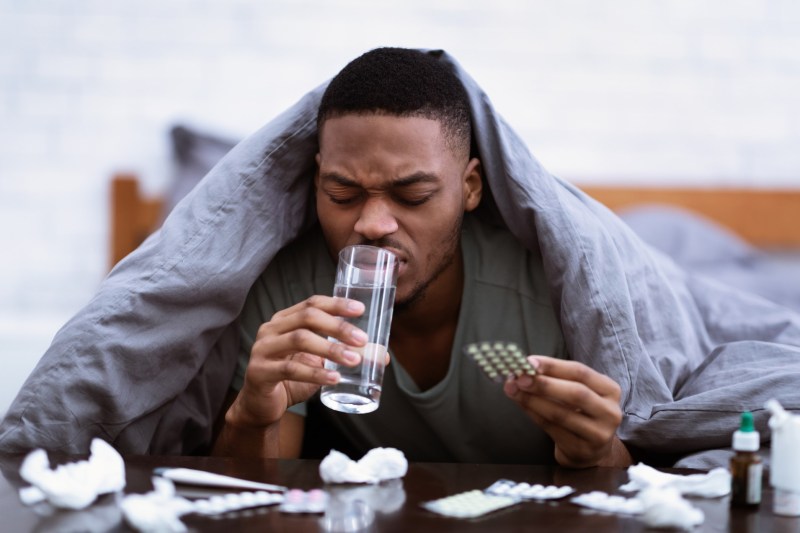 Sick African Man Taking Pills Drinking Water Lying In Bedroom.
