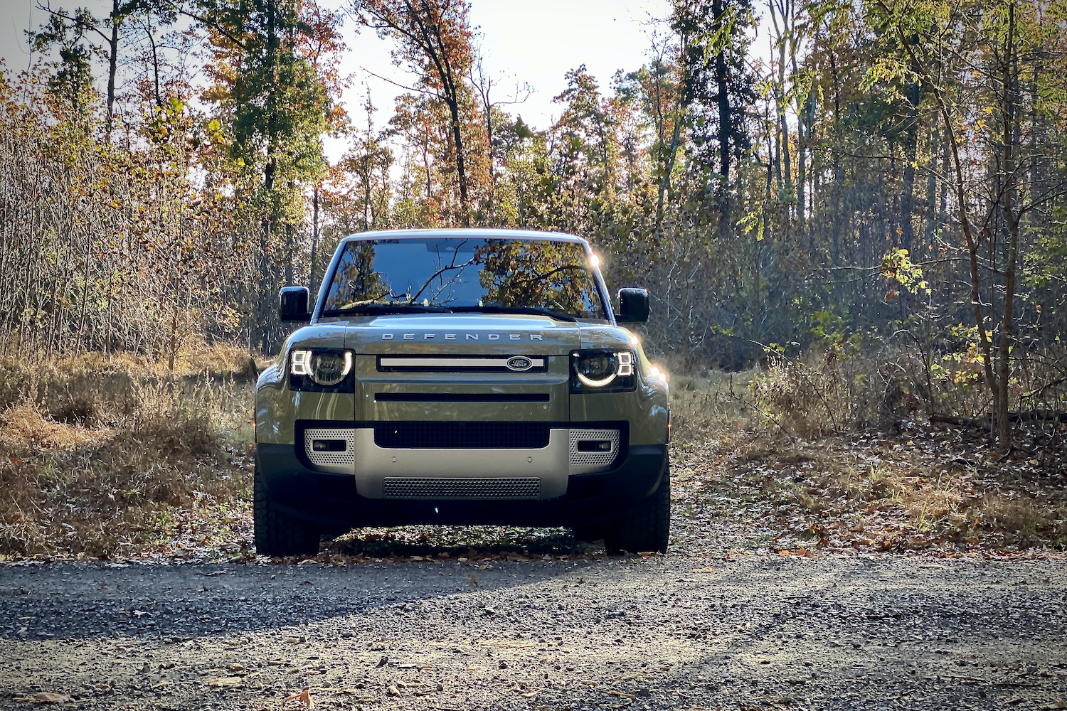 Close up of Land Rover Defender front end with trees in the back and a gravel road.