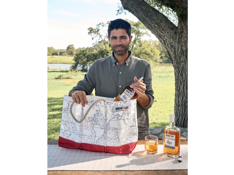 Adrian Grenier standing outside holding a bottle of Mount Gay Rum and a Mount Gay Rum tote.