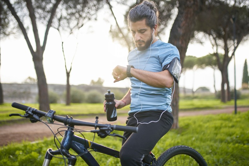 A male cyclist with water bottle checking his watch in a park on a sunny day.