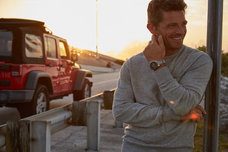 A man in a gray long-sleeved shirt wearing a luxury watch near a truck in the outdoors during a sunset. 