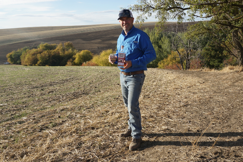 Farmer Nathan in the field holding a bottle of Copperworks Distilling's single malt, salmon-safe whiskey.
