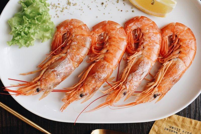 four cooked Prawns on a plate next to greens and a lemon wedge.