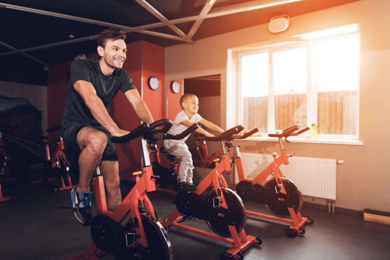 A father and his son using exercise bikes in a gym on a sunny day