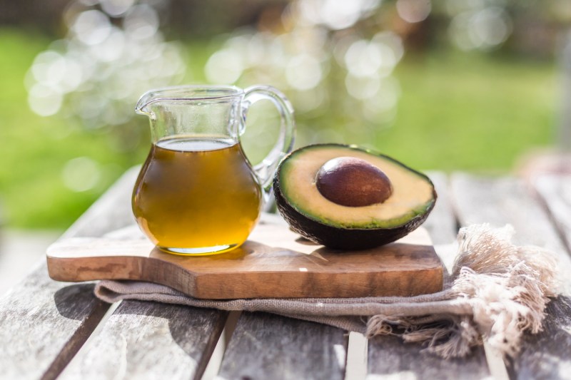 A pitcher of avocado oil beside a sliced avocado on a wooden board.