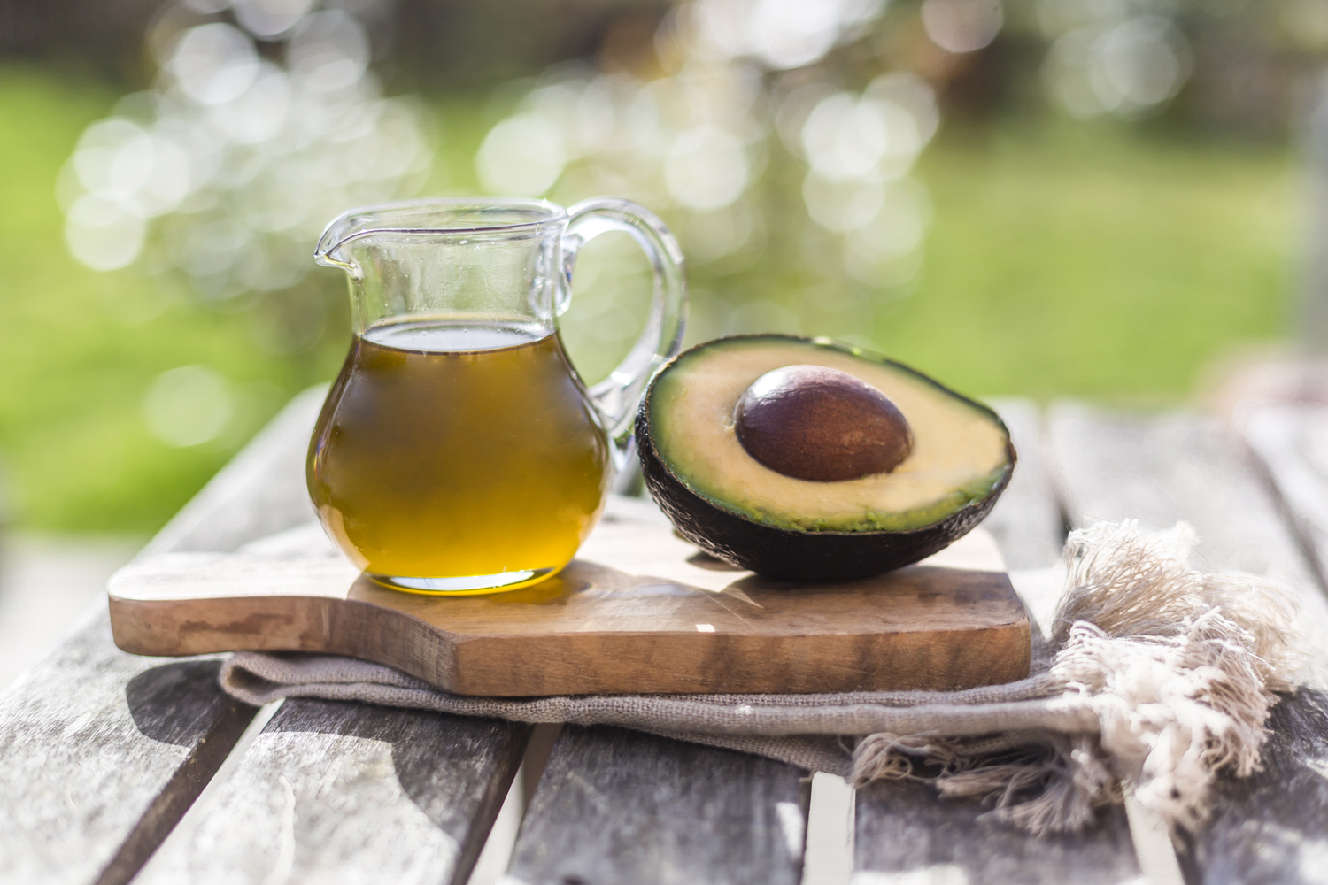 A pitcher of avocado oil beside a sliced avocado on a wooden board.