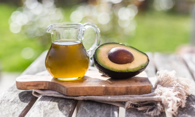 A pitcher of avocado oil beside a sliced avocado on a wooden board.