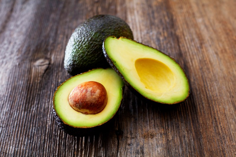 A whole avocado behind a sliced avocado on a wooden surface.
