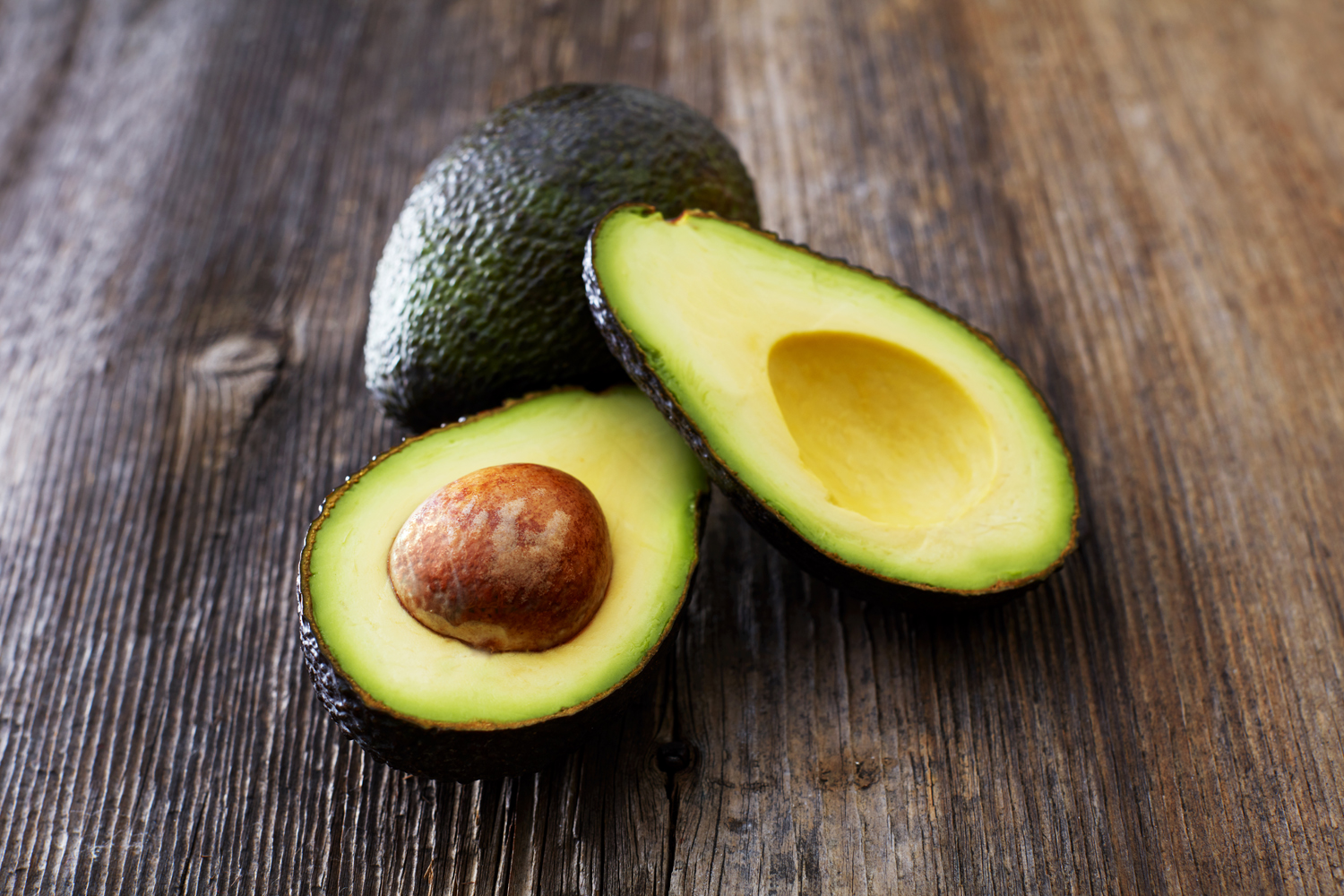 A whole avocado behind a sliced avocado on a wooden surface.