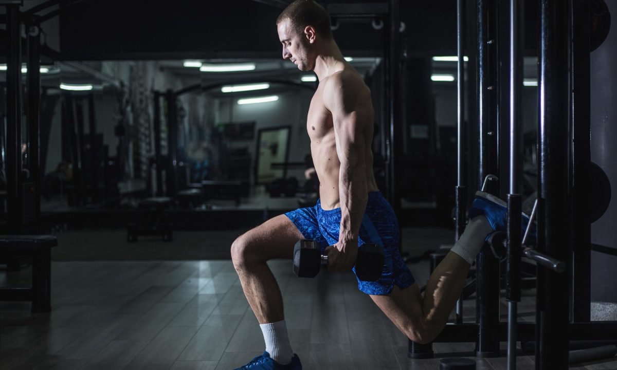 A shirtless man doing split squats using a dumbbell