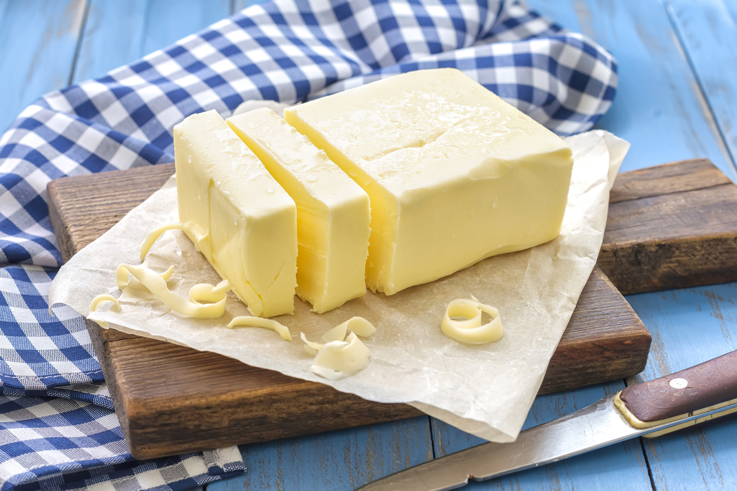Sliced butter on paper atop of a wooden cutting board with a knife and a blue and white checkered kitchen towel.