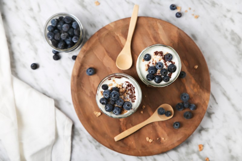 Dwie filiżanki parfait jogurt z jagodami i łyżki na okrągłej tacy obok miski jagód na stole.
