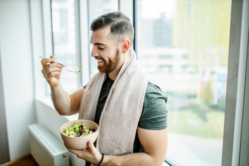 A man with a towel draped around his neck eating salad by the window.