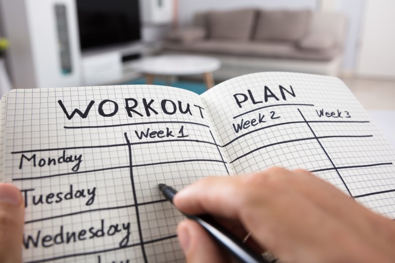 A person's hand writing out their workout plan on a notebook in a room.