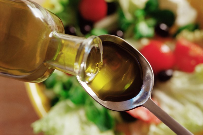 Olive oil being poured onto a spoon, with a bowl of salad in the background