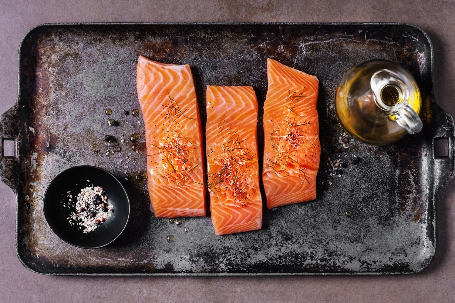 Three salmon slices with a pitcher of olive oil and a bowl of spices on a black tray.