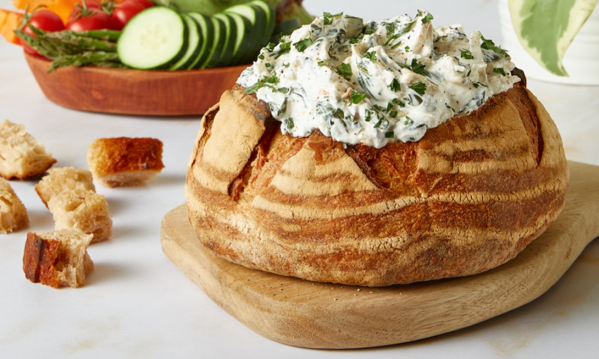 Good Culture Spinach and Artichoke dip placed in a bread bowl on a white kitchen counter with salad in the back.