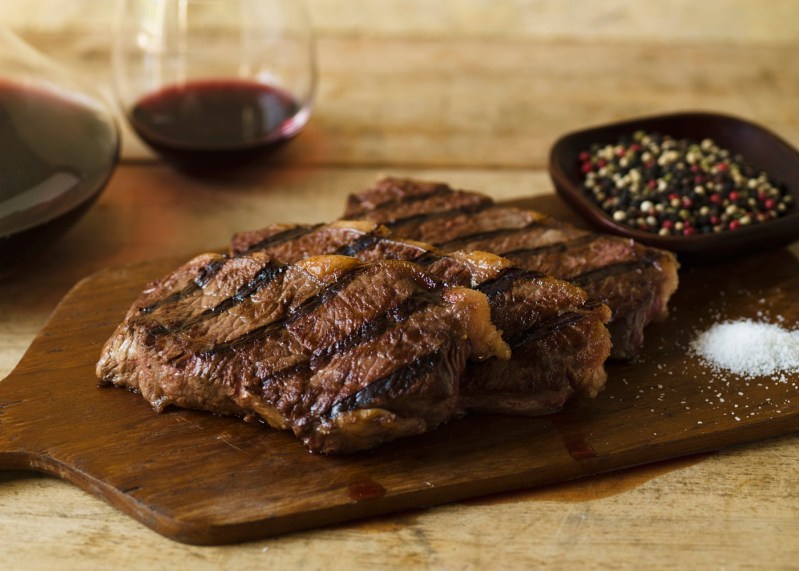 Slices of steak, a bowl of peppercorn, and salt mound on a wooden cutting board