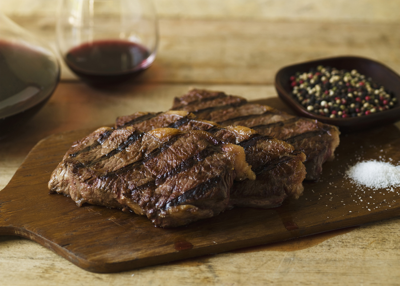 Slices of steak, a bowl of peppercorn, and salt mound on a wooden cutting board