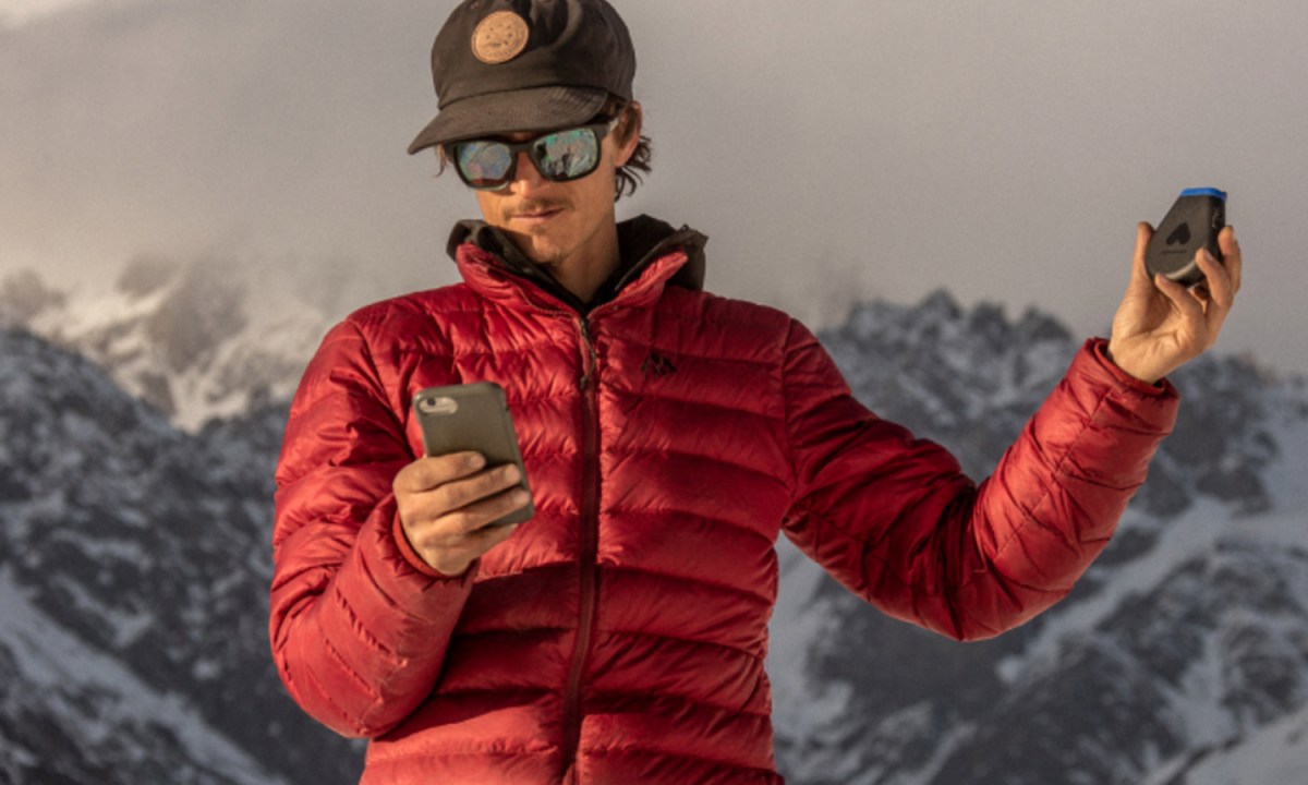 A man holding a phone in his right hand and a Somewear Global Hotspot in his left hand in the mountain.