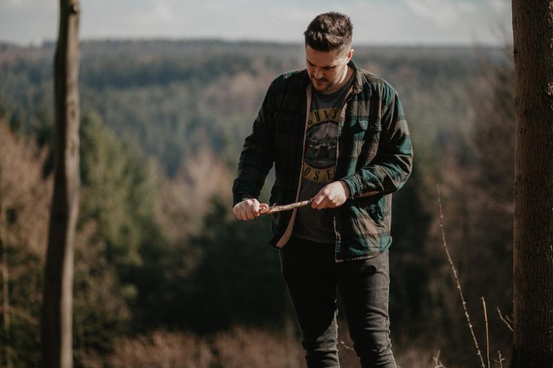 A man in a green and gray jacket whittles a stick to a sharp point