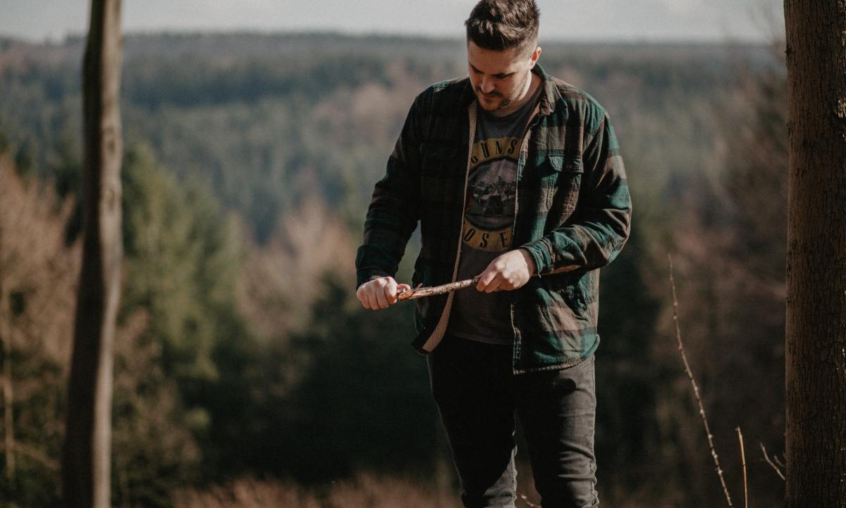 A man in a green and gray jacket whittles a stick to a sharp point