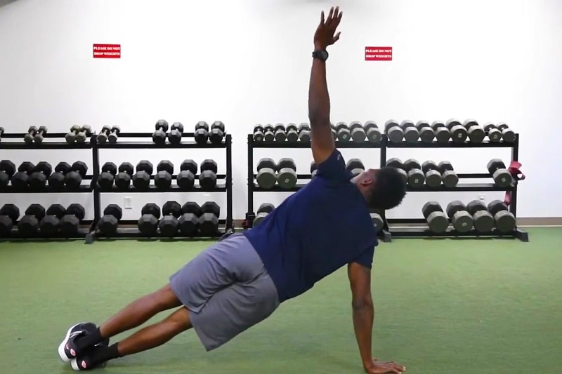 A male athlete doing thoracic rotations with dumbbells on racks in the background.