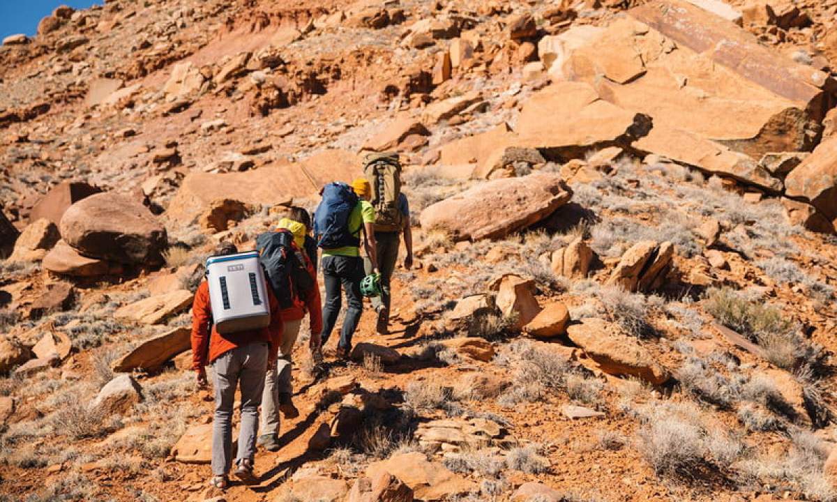 Four people hiking on a rocky trail. The last member is carrying a Yeti backpack cooler.