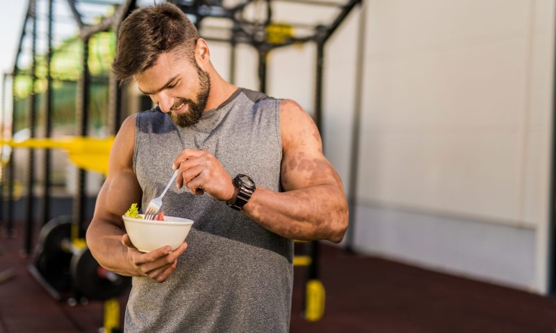 A man in gray sleeveless top eating a vegetable salad in the gym.