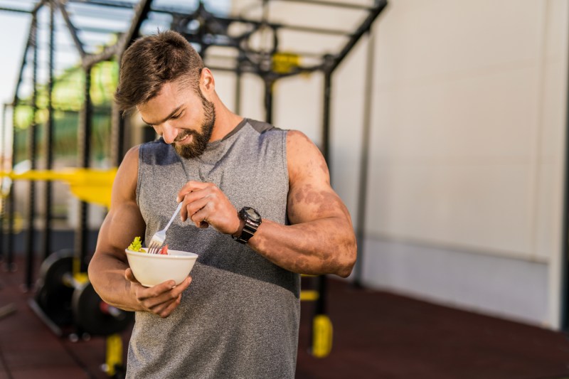 Un hombre con top gris sin mangas comiendo una ensalada de verduras en el gimnasio.