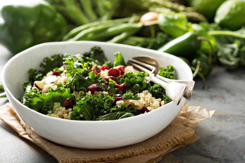 A bow of kale and quinoa salad on brown cloth with vegetables in the background.