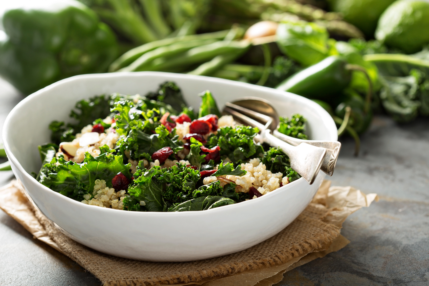 A bow of kale and quinoa salad on brown cloth with vegetables in the background.