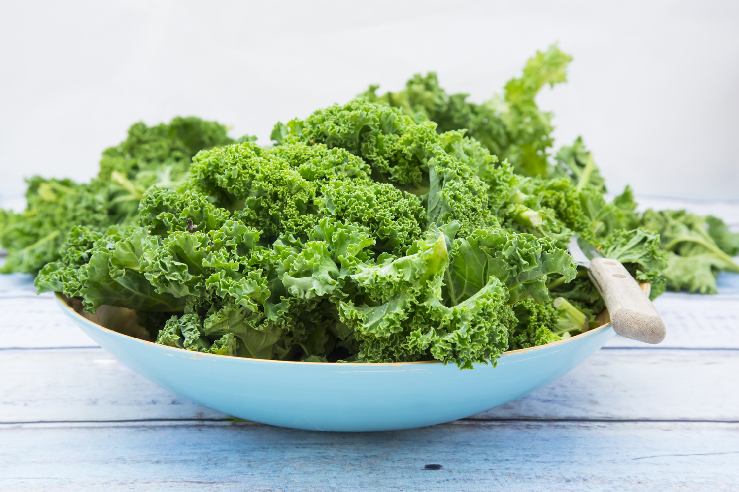 Fresh kale leaves and a knife on a white bowl on a white wooden table.