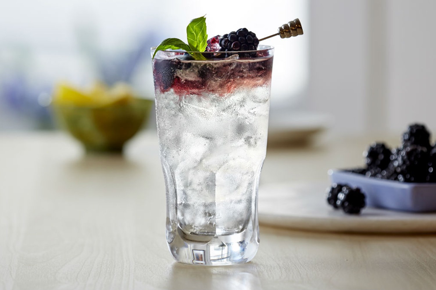A Bacardi Blackberry Bramble garnished with basil and blackberry skewer with a tray of blueberries in the background.
