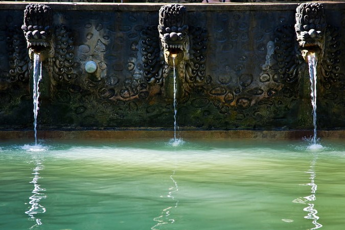 A green hot springs pool with three animal heads over it. There seems to be water pouring out of the animal heads' mouths.