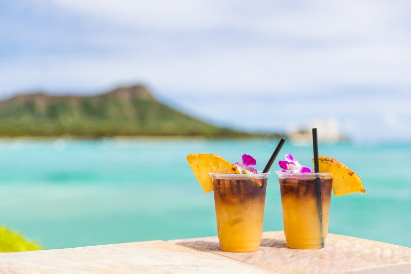 Two Mai Tai cocktails topped with flowers and pineapples on a surface with a view of the beach