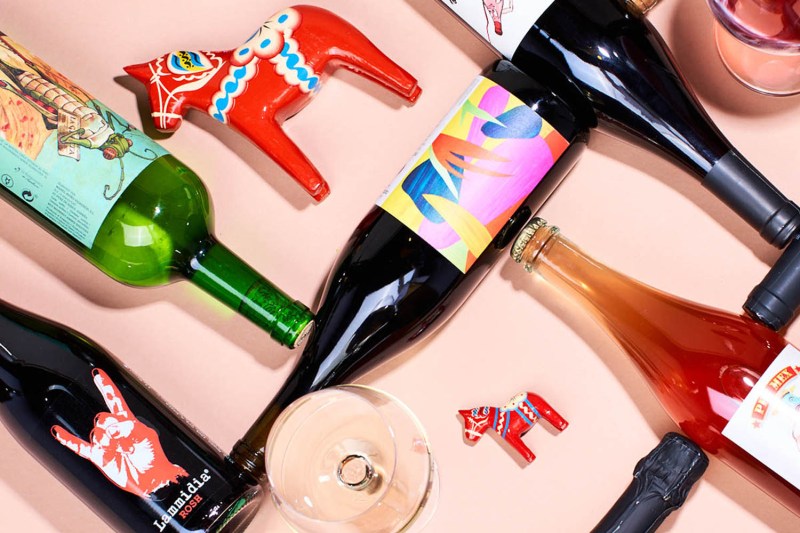 An overhead shot of an empty wine glass, a glass filled with wine, wine bottles, and differently sized red wooden horses on pink table.