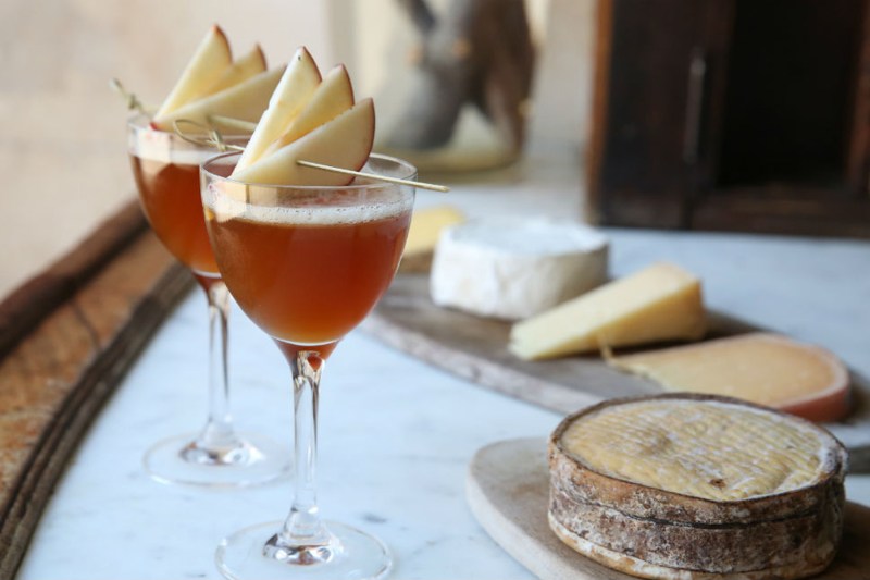 Two glasses of Between the Apple Trees cocktails garnished with apple slices beside cheeses on oval-shaped wooden plates.