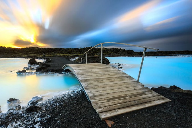 A large blue hot spring with a brown bridge over it. The sky is yellow, grey, and blue.