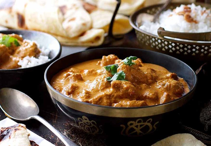A butter chicken served in a bowl beside a metal spoon with various dishes in the background.