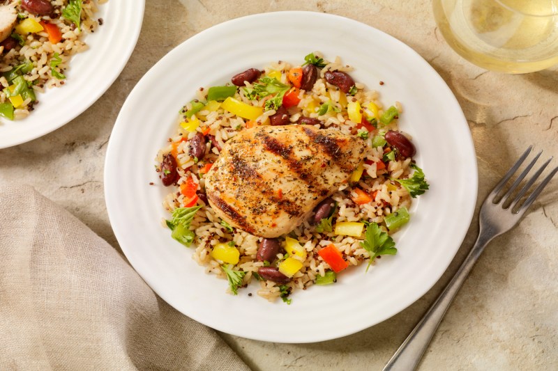 An overhead shot of grilled chicken with rice and vegetables beside a metal fork and a glass of wine on a table.