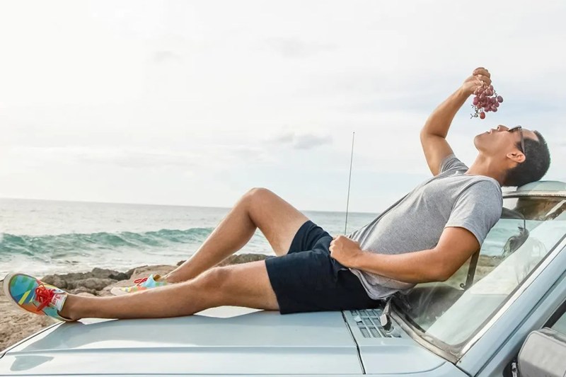 Man in Chubbies Shorts laying on the hood of a car while holding a grape over his mouth.